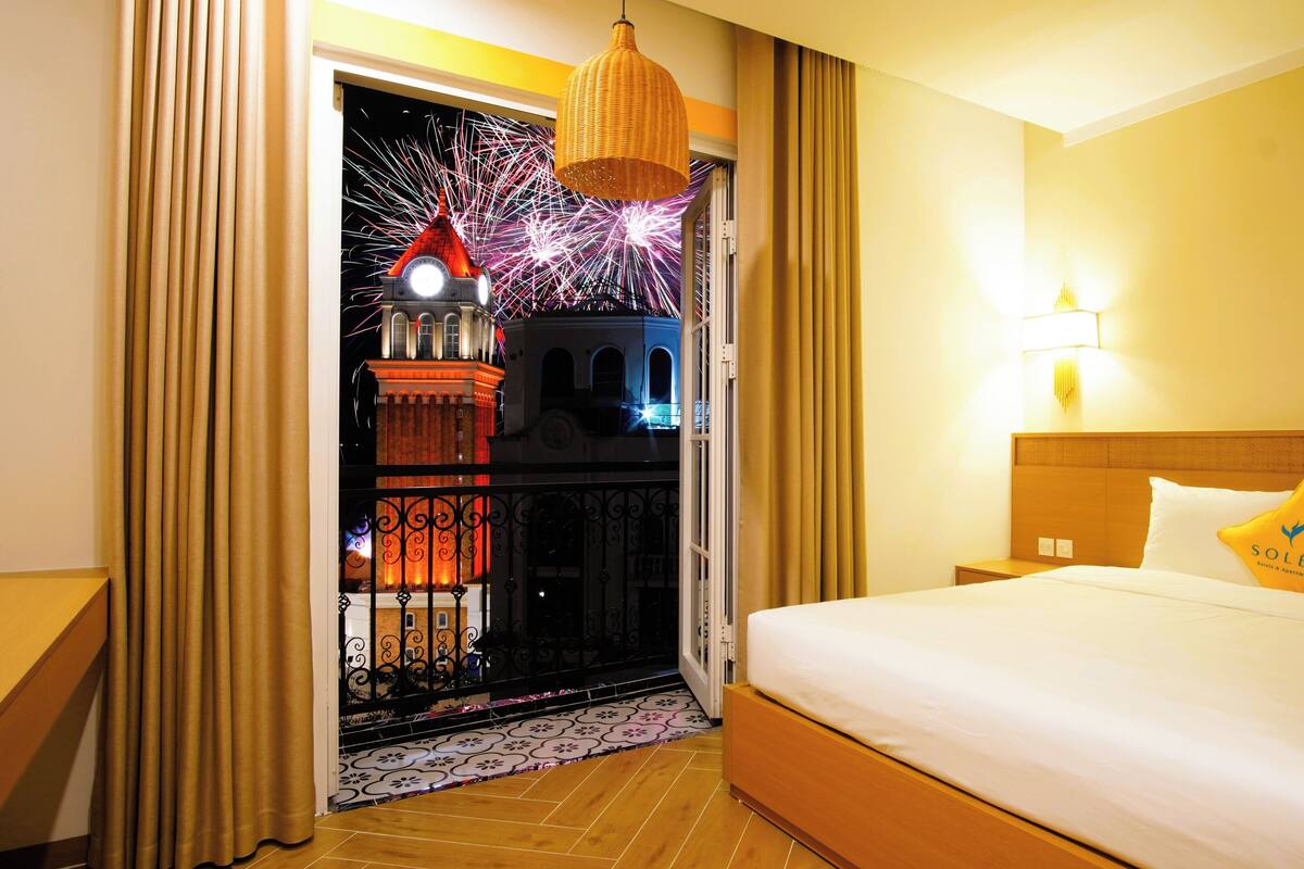 Une chambre d’hôtel avec un lit, une table de chevet, une lampe et une vue sur un immeuble illuminé par des feux d’artifice.