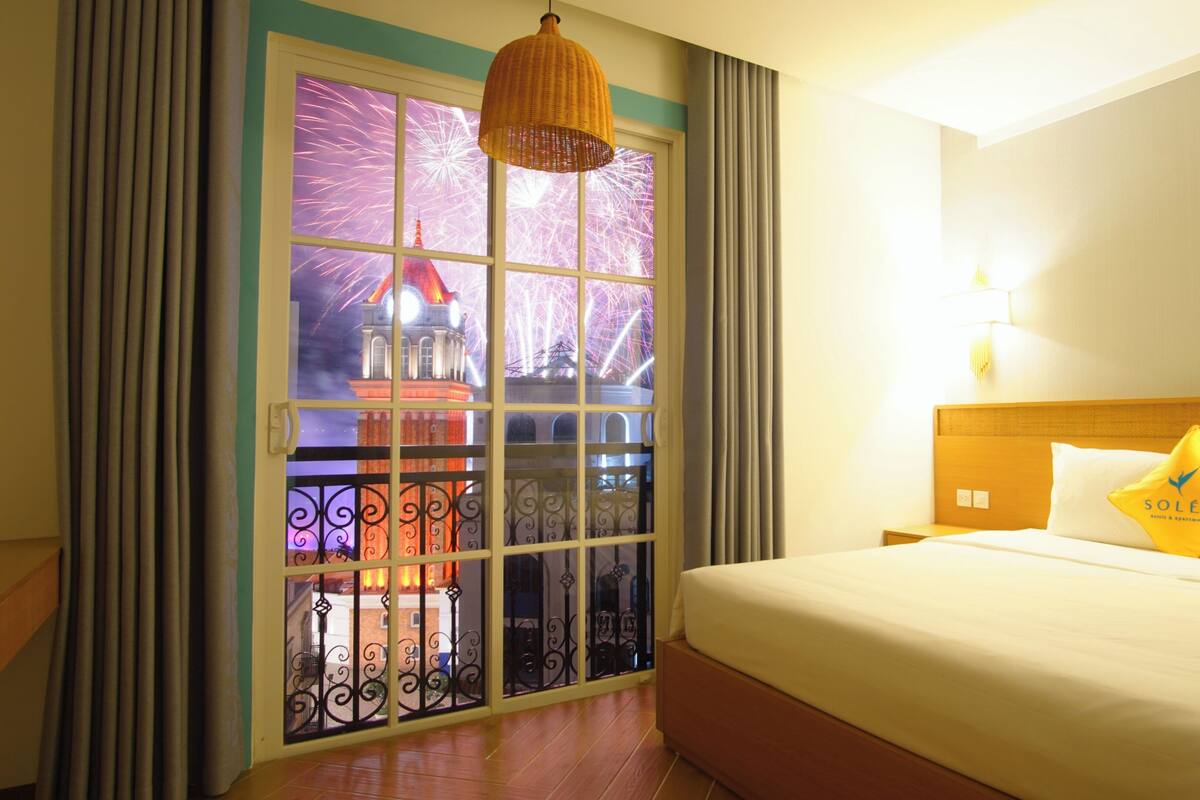Une chambre d’hôtel avec un grand lit, une fenêtre donnant sur un spectacle de feux d’artifice et un balcon agrémenté d’une grille en fer forgé.