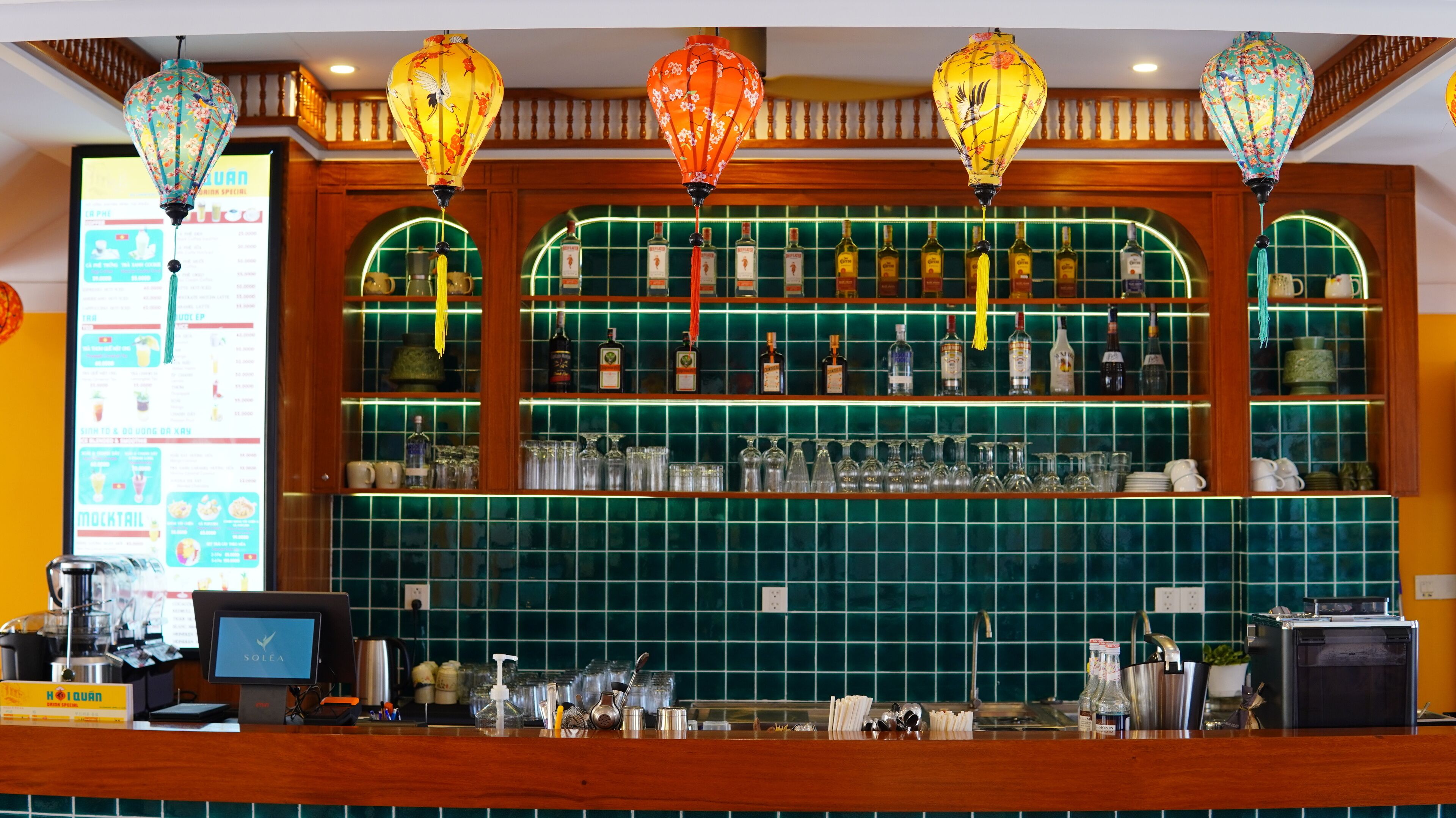 Un espace bar agrémenté d’un mur recouvert de carreaux verts, d’étagères en bois où sont exposées des bouteilles d’alcool et de lanternes décoratives.