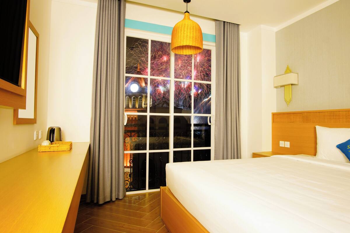 Une chambre d’hôtel avec un lit, une table de chevet, une lampe, une fenêtre avec des rideaux et une vue sur les feux d’artifice.