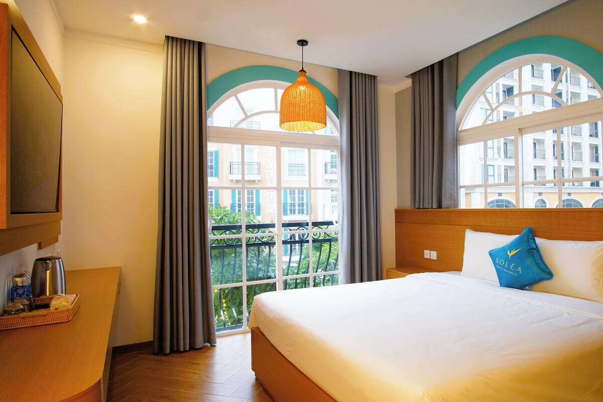 Une chambre d’hôtel avec un grand lit, une tête de lit en bois, une table de chevet, une télévision et une fenêtre avec des rideaux.