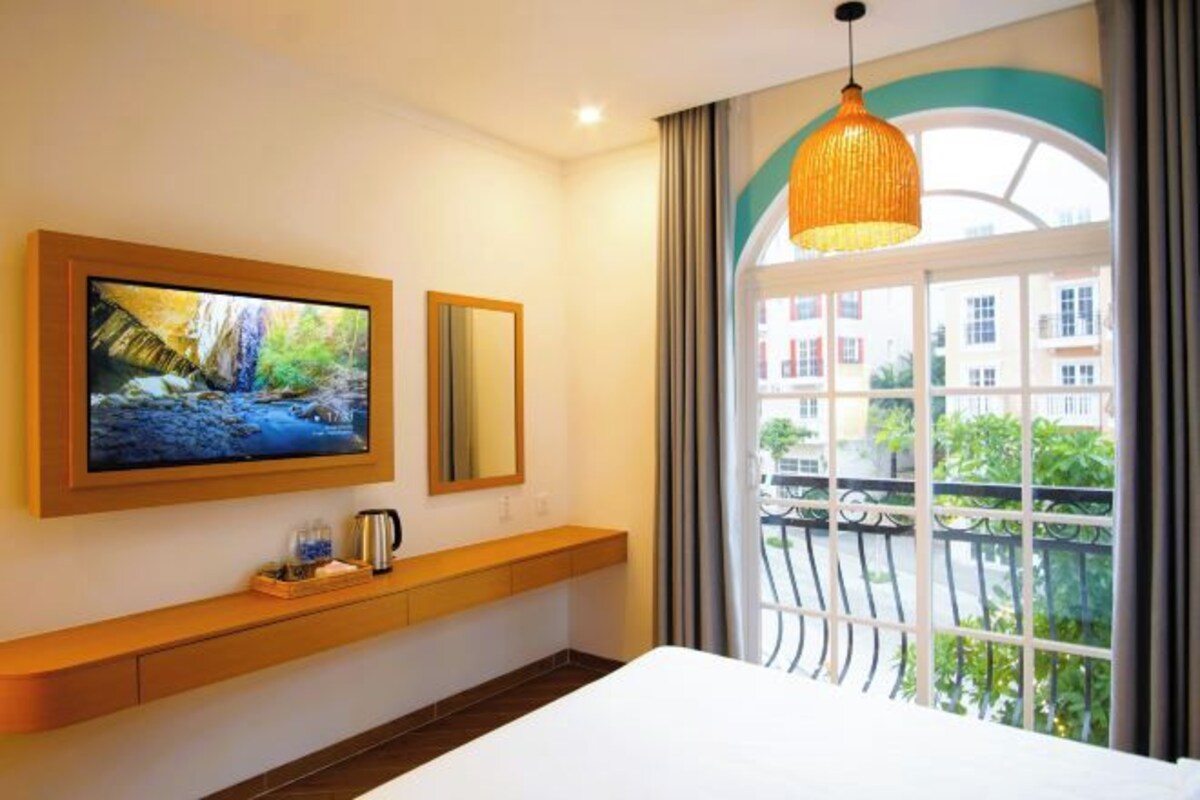 Une chambre d’hôtel avec un grand téléviseur fixé au mur, une table de chevet avec une bouilloire, et une vue sur les bâtiments à travers une fenêtre donnant sur un balcon.
