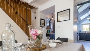 A dining area with a table set for two, featuring clear glassware, a pitcher, and a vase with flowers. A staircase with wooden railings is visible in the background.