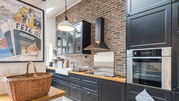 A modern kitchen with a brick wall, black cabinets, stainless steel appliances, and a hanging light fixture.