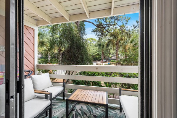 A balcony with a white railing, two white cushioned chairs, a small wooden table, and a view of trees and a road.