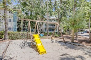 Ein Spielplatz mit Rutsche und Schaukeln, umgeben von Bäumen und einem Gebäude im Hintergrund.
