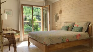 Un dormitorio con una cama de madera, una silla de mimbre, una mesita, un espejo y vistas a zonas verdes a través de un ventanal.