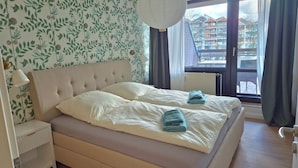 A bedroom with a large bed, a nightstand, a hanging light fixture, and a view of a building through a window. - 5 DTV star vacation apartment on Lake Stella with marina view (Bad Zwischenahn)