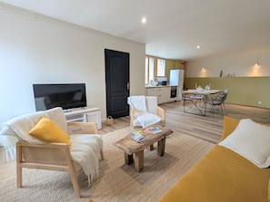 Ein Wohnzimmer mit Fernseher, einer Couch, Sesseln, einem Couchtisch und einer Küche im Hintergrund.