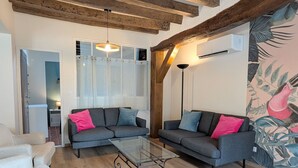 A living room with two grey sofas, a glass coffee table, wooden beams, a ceiling light, and a wall with a tropical mural.