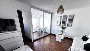 A modern living room with a TV, white furniture, a dining area with a table and chairs, and a large sliding glass door leading to a balcony. - Quiet Studio Cabin, Canal View, Near Beach and Shops (Cap d'Agde)