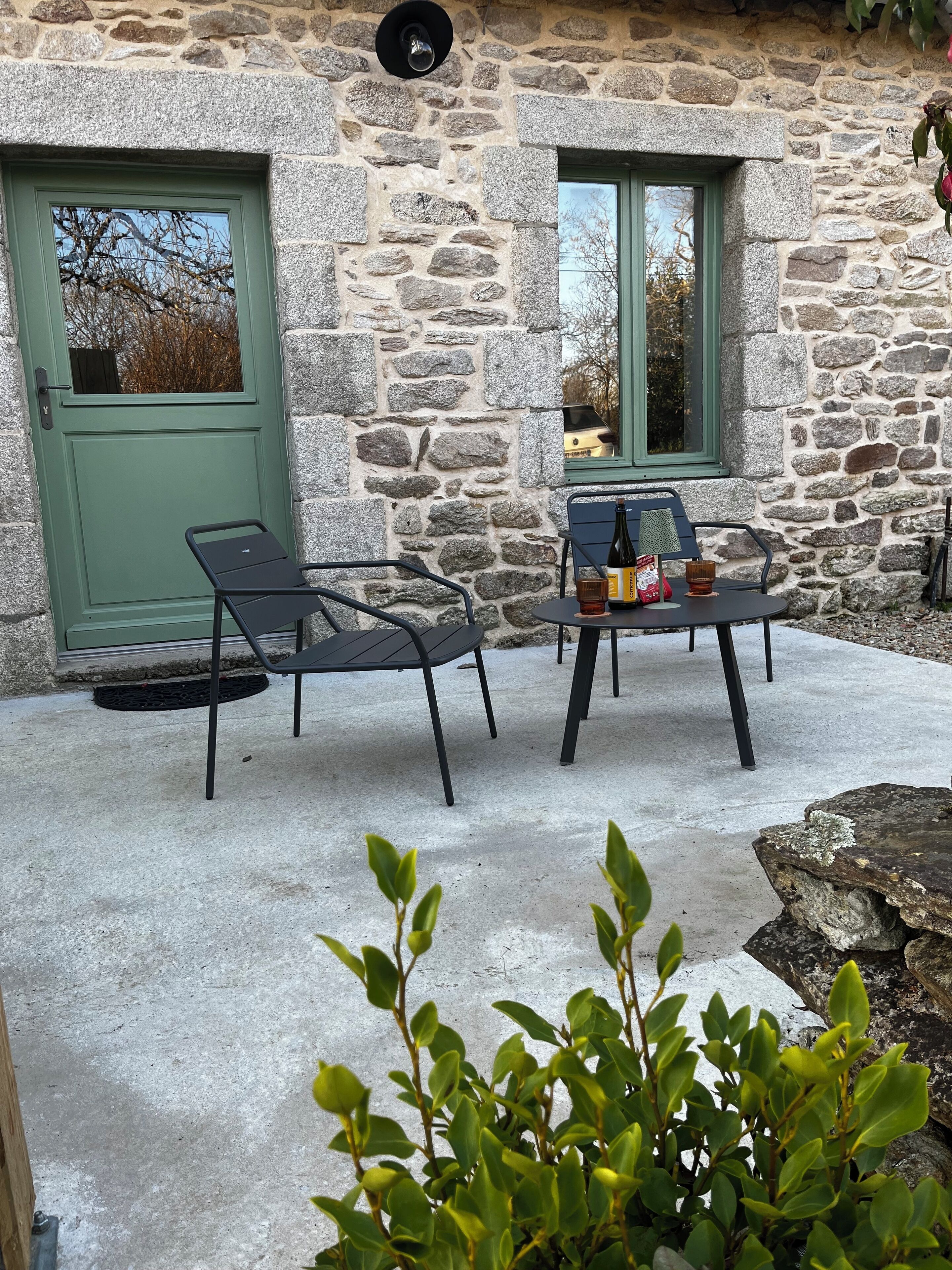 A stone building with a green door and window, outdoor seating with chairs and a table, and a small plant in the foreground.