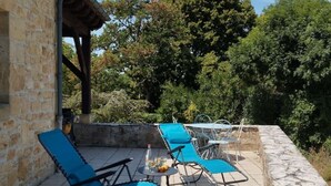 Un patio exterior con paredes y piso de piedra, y vista a árboles. Hay sillones azules y una pequeña mesa redonda con una botella y vasos.