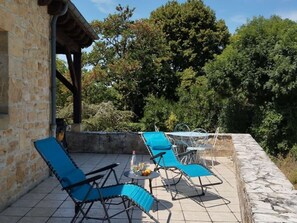 Eine Terrasse im Freien mit Steinmauern, Steinboden und Blick auf Bäume. Blaue Lounge-Stühle und ein kleiner runder Tisch mit einer Flasche und Gläsern.