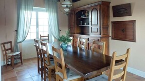 Comedor con mesa y sillas de madera, un armario con cristalería y una ventana con cortinas.