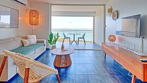 A modern living room with a sofa, a round coffee table, a wicker chair, a TV, and a balcony with a view.
