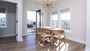 A dining area with a wooden table and chairs, a modern chandelier, and large windows with a view of buildings. - New Build-Rooftop-Steps from the Beach! Unit Salt (Virginia Beach)