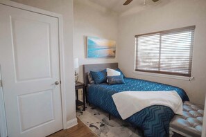 Une chambre à coucher comprenant un lit, une table de chevet, une lampe, une fenêtre avec des stores, une porte et un tableau accroché au mur.