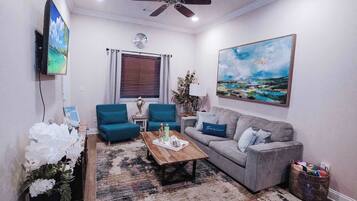 A living room with a sofa, a coffee table, a TV mounted on the wall, a ceiling fan, and a window with blinds.
