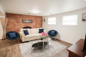 A living room with a brick fireplace, a white sofa with blue cushions, two blue chairs, a glass coffee table, and a TV stand. - Midwest Haven- Spacious, Family Home, Near Airport (Kansas City)