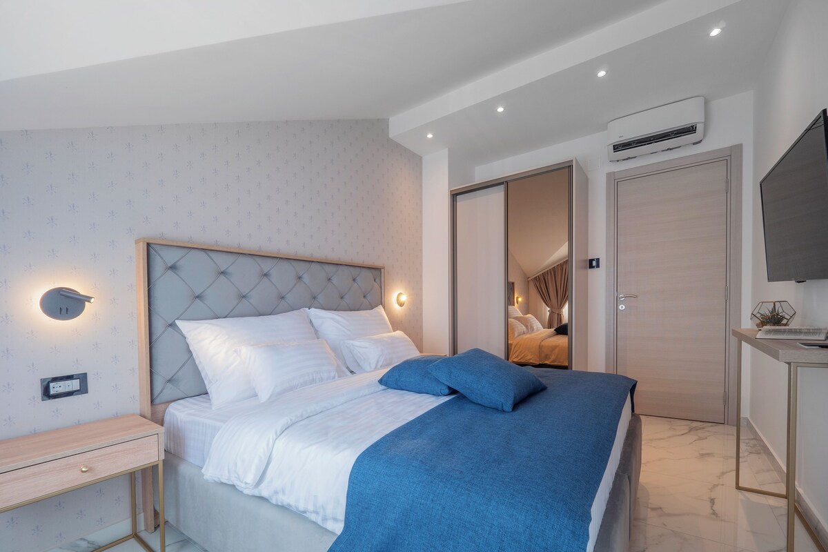 A modern bedroom with a tufted headboard, a bedside table, a bed with white bedding and a blue throw, an air conditioning unit, and a television on a stand.
