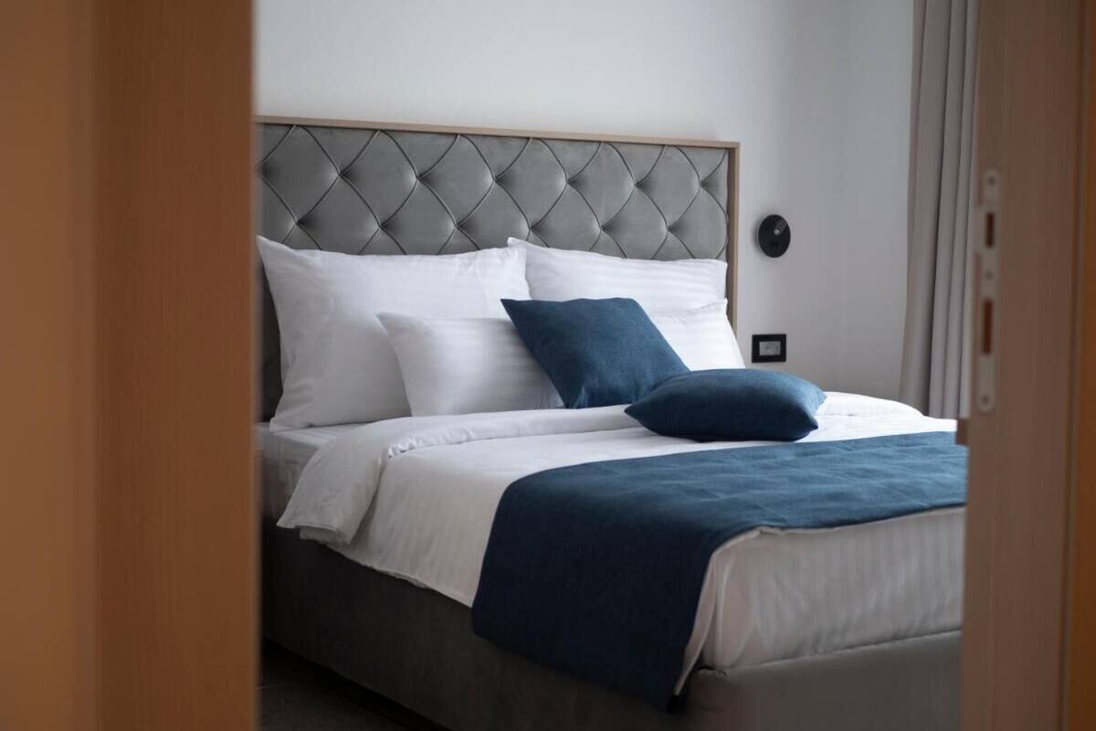 Una cama bien hecha con cabecera capitonada, ropa de cama blanca y azul, y una manta azul oscuro.