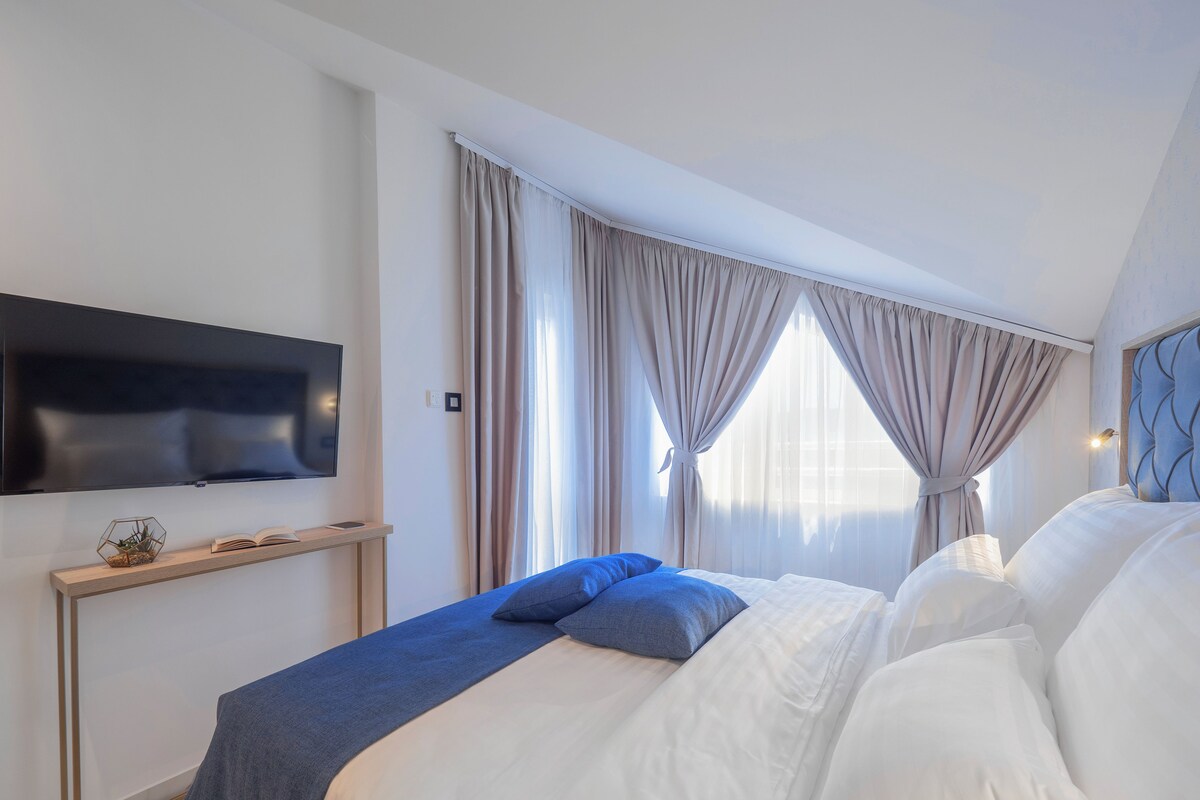 A modern hotel room with a flat-screen TV, a bedside table with a decorative item, a bed with white and blue bedding, and large windows with curtains.