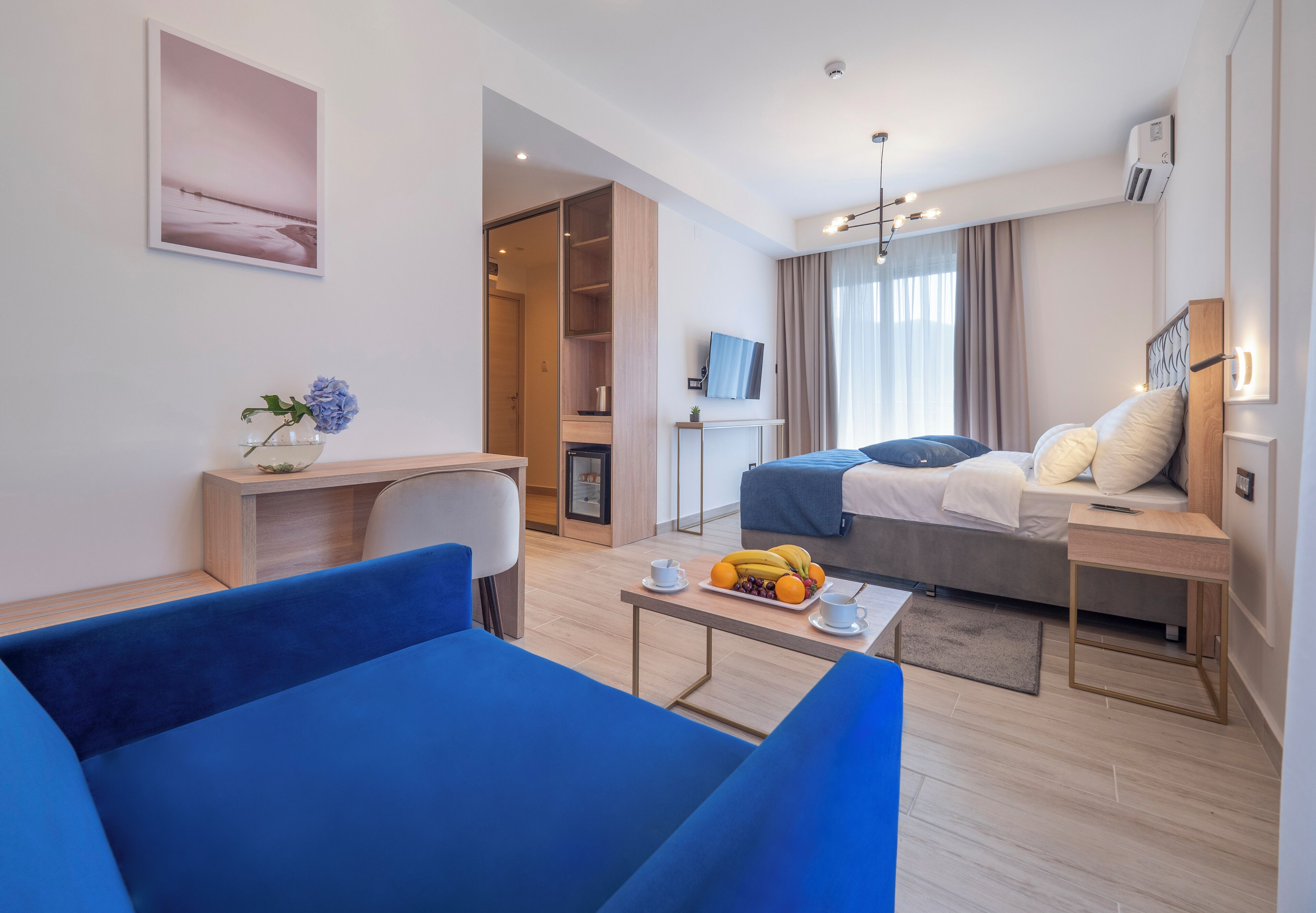 Una habitación de hotel moderna con una cama, un escritorio, un sofá azul, una mesita con fruta y un televisor.
