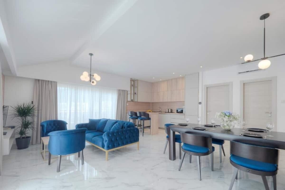 Una sala de estar moderna con un sofá azul, un comedor con una mesa de mármol y una cocina al fondo.