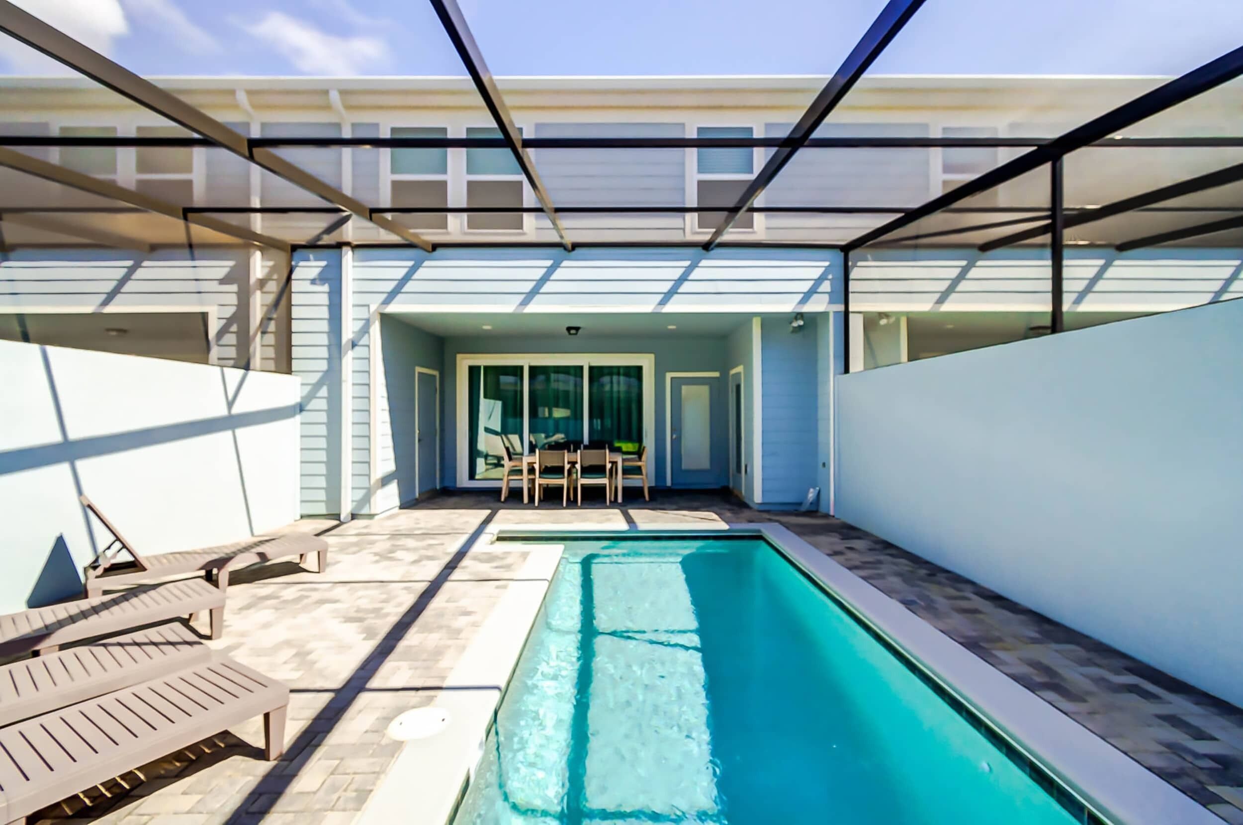 A swimming pool with lounge chairs and a table, enclosed patio area with glass doors, and a view of the sky.