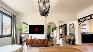 A spacious living room with a large flat-screen TV, wooden furniture, and a chandelier.