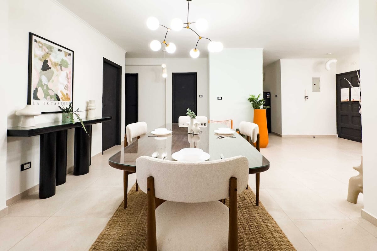Ein moderner Essbereich mit einem Glastisch, weißen Stühlen und einer zentralen Leuchte.