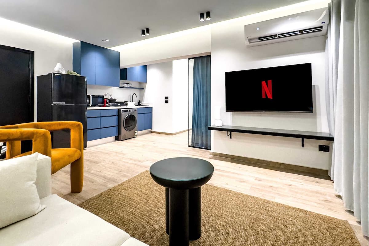 A modern living room with a sofa, a coffee table, a TV mounted on the wall, and a kitchen in the background.
