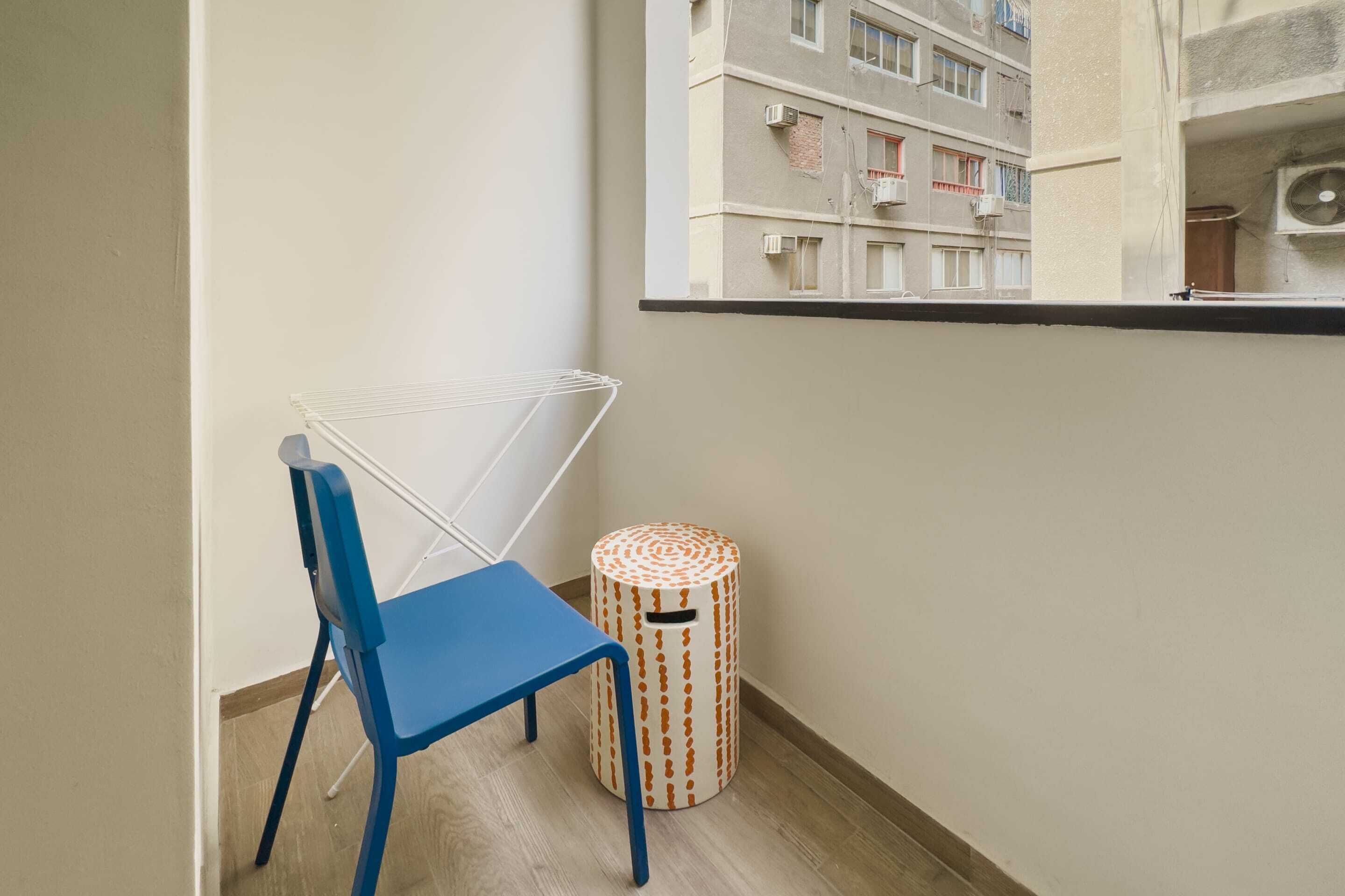 A corner of a room with a blue chair, a white drying rack, and a patterned trash bin.