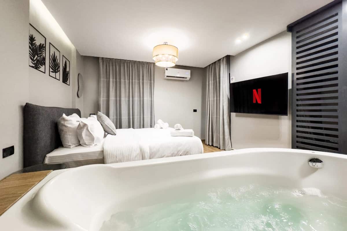 A modern hotel room with a large white bathtub, a bed with white linens, a ceiling light, and a Netflix logo on the wall.