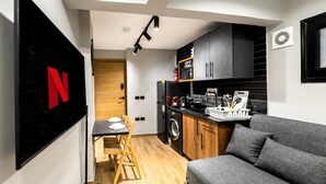 A compact living space with a kitchenette, a grey sofa, a dining table with chairs, and a TV mounted on the wall. - Xuru Stays I Heliopolis 1 - 5 Min to Airport (Cairo)