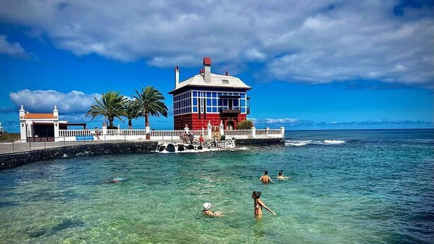 Une mer d’un bleu profond où l’on voit des gens nager, et un bâtiment à la façade rouge et blanche.