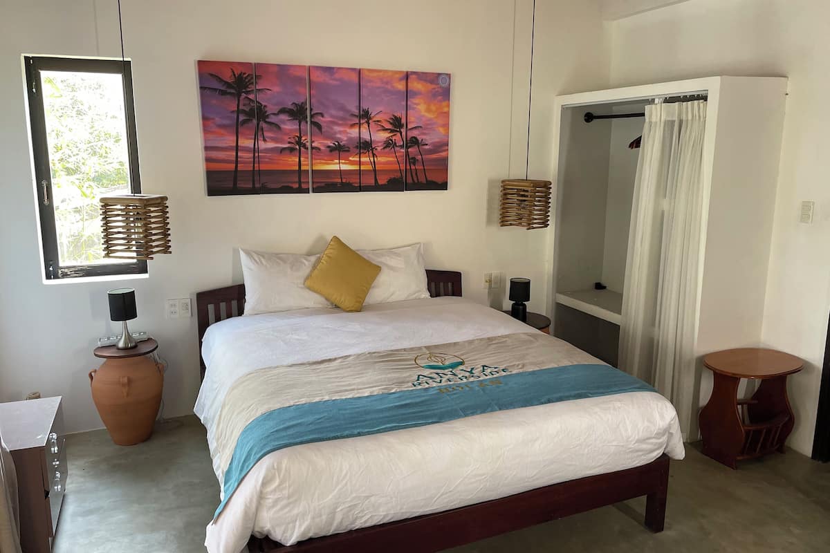 A bedroom with a bed, a large painting of a sunset, a window with a curtain, and a small table with a lamp.
