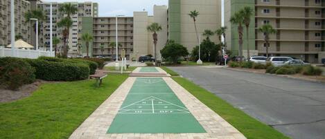 A courtyard with a shuffleboard court, surrounded by a fence, palm trees, and a parking area with cars.