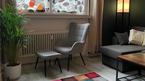 Ein modernes Wohnzimmer mit grauem Sofa, einem kleinen Tisch, einem Sessel, einem farbenfrohen Teppich und einem Fenster mit gemusterten Vorhängen.