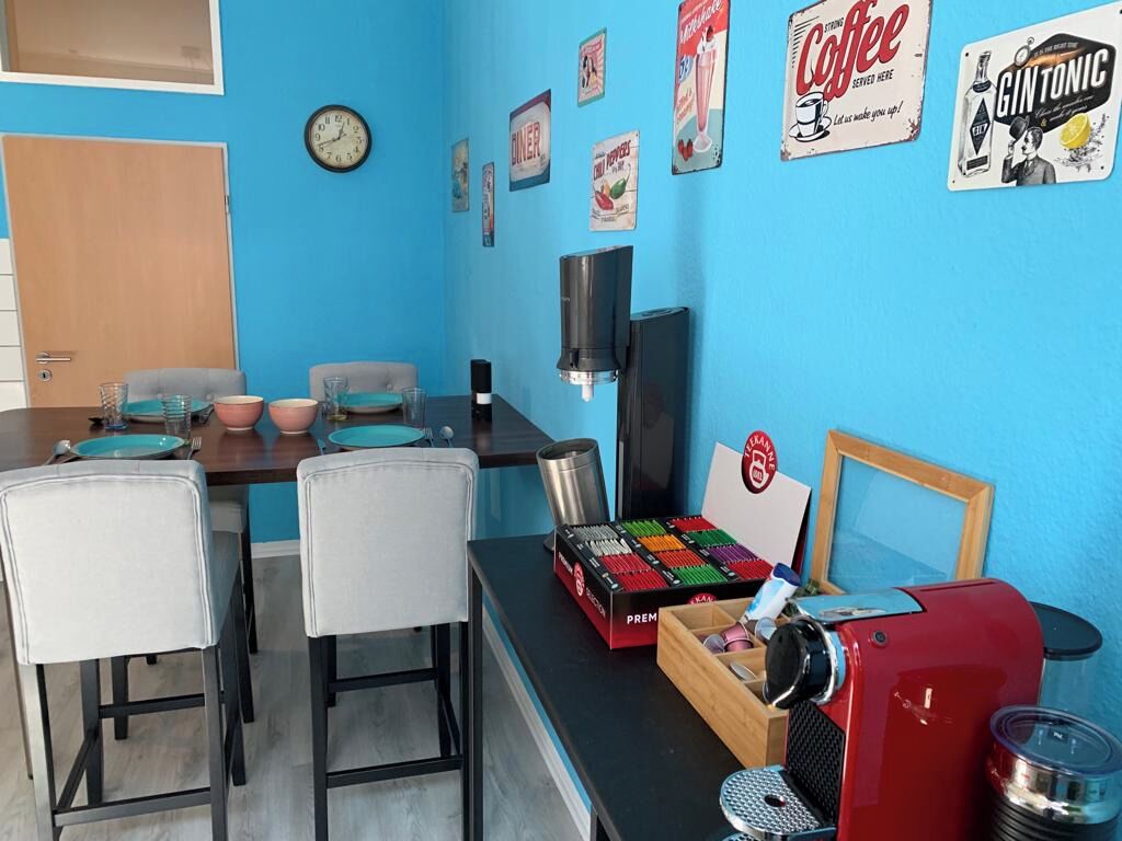 Een kleine, vrolijk gekleurde kamer met een blauwe wand, een houten deur en een tafel gedekt met borden en glazen. Op de wand hangen vintage borden met daarop koffie en gin-tonic, en op de tafel staat een rode koffiemachine.