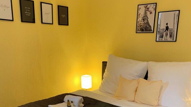 Ein Hotelzimmer mit einem Bett, Kissen, einem Nachttisch und einer Lampe. An der Wand hängen gerahmte Bilder.