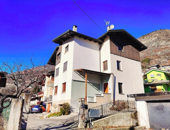 A two-story building with a balcony, a car parked in front, and a mountainous backdrop.