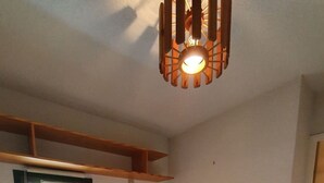 Ein kleines Zimmer mit einem Einzelbett, einem Holzbettgestell, einem kleinen Nachttisch, einem Fenster und einer Deckenleuchte.