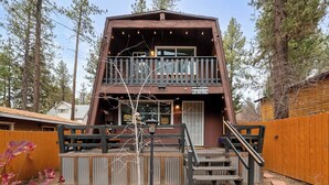A two-story wooden cabin with a balcony, a wooden deck, and a staircase leading to the upper level. - Moonridge Manor Cozy Retreat with Hottub (Big Bear Lake)