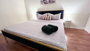 A neatly made bed with a floral-patterned bedspread and two rolled towels, a bedside table with a lamp, and a wall-mounted abstract painting. - Nice 1-bedroom apartment in Marina del Rey (Marina del Rey)