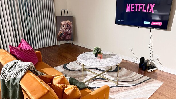 A modern living room with a large flat-screen TV displaying 'NETFLIX', a round marble coffee table, an orange sofa with patterned cushions, and a decorative owl painting. - Nice 1-bedroom apartment in Marina del Rey (Marina del Rey)