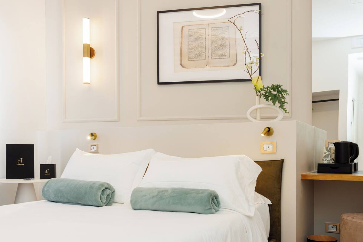 Un lit bien fait, avec du linge de lit blanc, une table de chevet avec une lampe et un livre encadré accroché au mur.