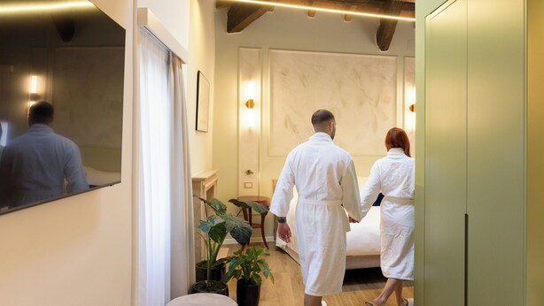 A hotel room with a bed, a mirror, a potted plant, and a wooden floor.