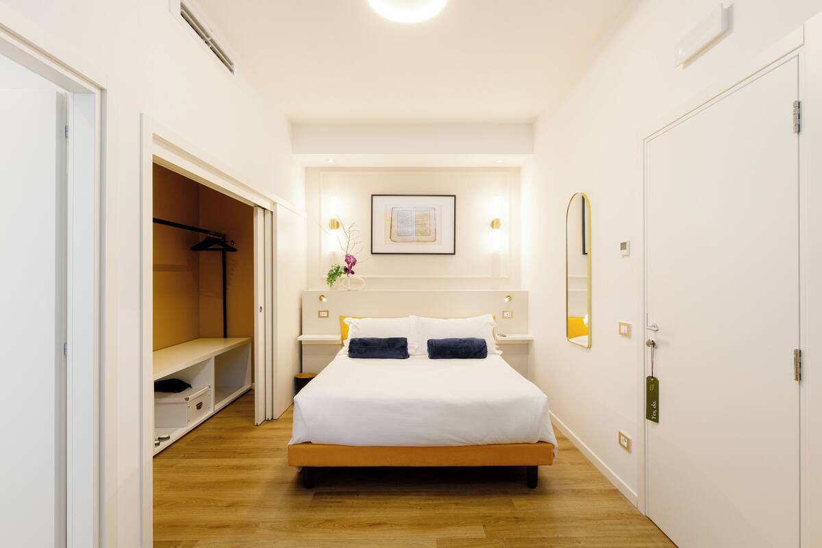 Une chambre à coucher comprenant un lit, une table de chevet, une commode, un miroir, un tableau encadré au mur et un placard avec des vêtements suspendus.